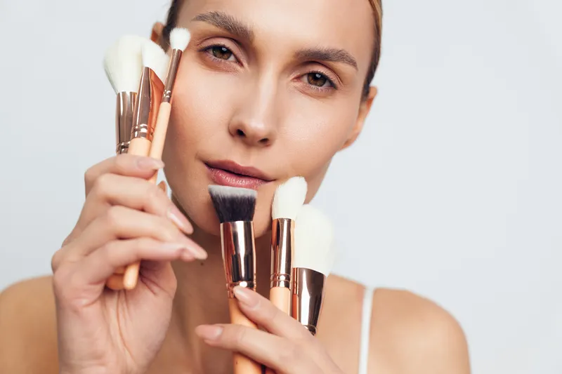 Young woman holds various makeup brushes close to her face with natural skin tone and neutral expression isolated on white background for beauty concept.