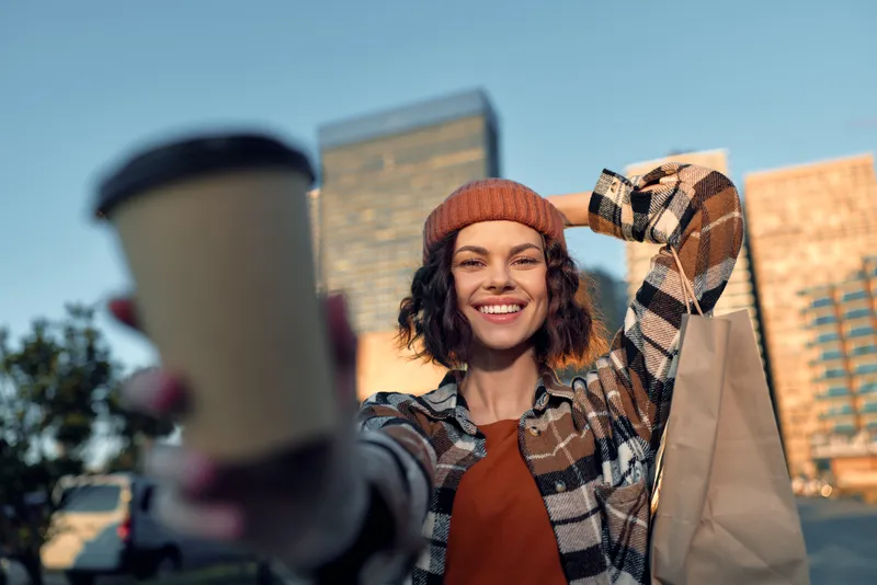 Woman holding coffee and shopping bag in urban street, smile toward camera during golden hour glow; candid lifestyle portrait with authenticity, mindful living and emotional storytelling