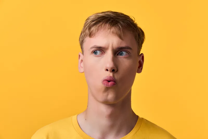 Man with a puzzled expression and pursed lips against a bright yellow background, showcasing curiosity and playful thinking in a casual sweater for lifestyle or reaction imagery
