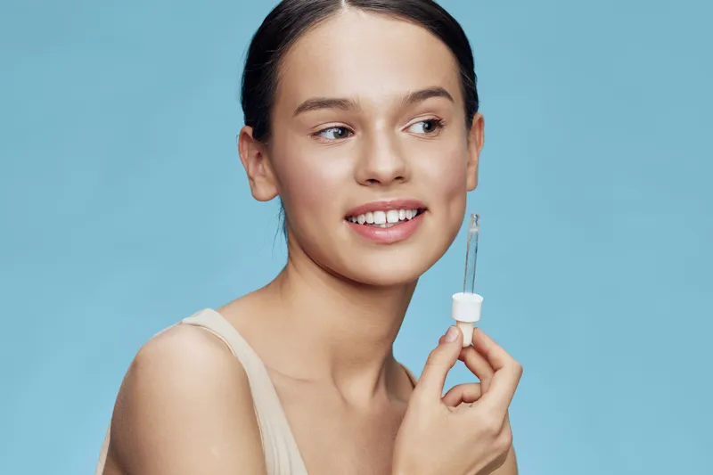 Young woman holding serum dropper, smiling against a blue background, showcasing skincare routine and healthy skin concept