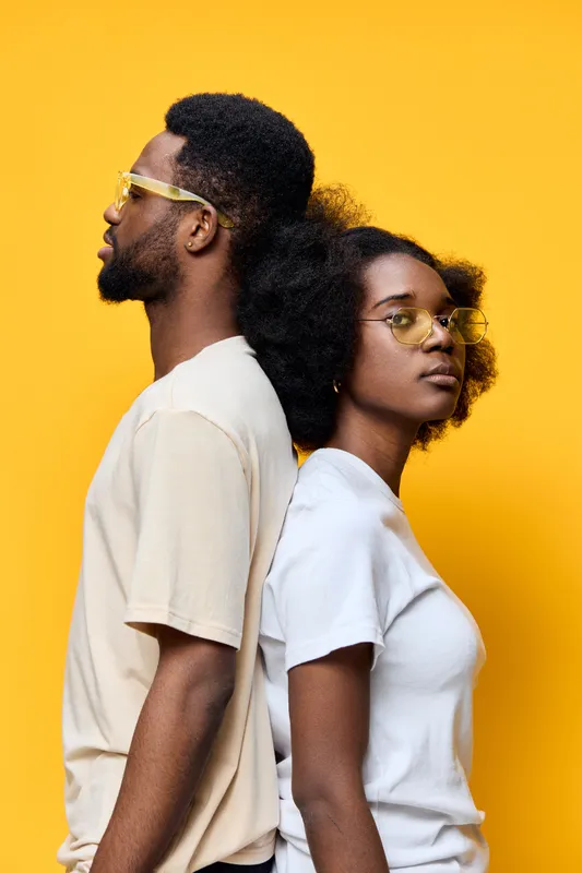 Stylish African American couple wearing trendy sunglasses, posing back to back against a vibrant yellow background, conveying confidence and fashion forwardness