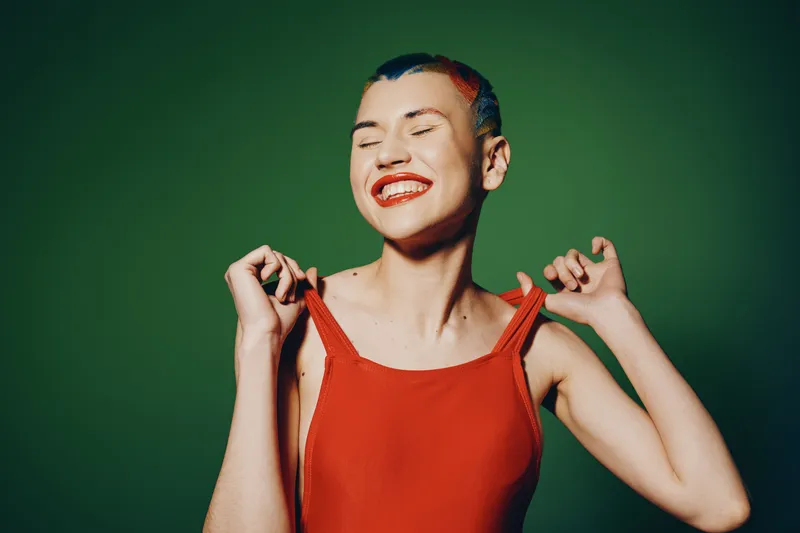 joyful woman with colorful hair and bright red lipstick wearing a red tank top smiling with closed eyes against green background