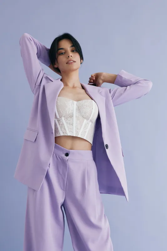 Stylish woman in lilac suit posing confidently against pastel background wearing white lace corset top, modern fashion look with soft color palette and casual elegance.
