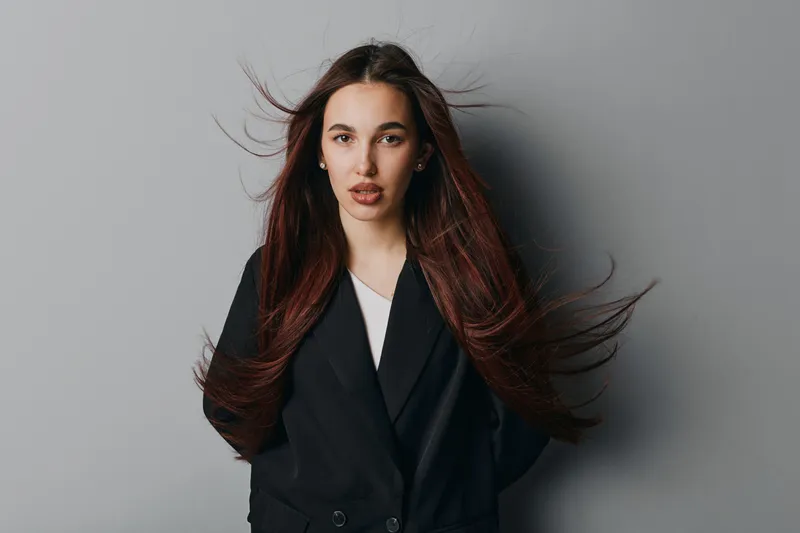 Young woman with long brown hair and serious expression wearing black blazer standing against gray background. Studio portrait of confident female model with natural makeup and flowing hair.
