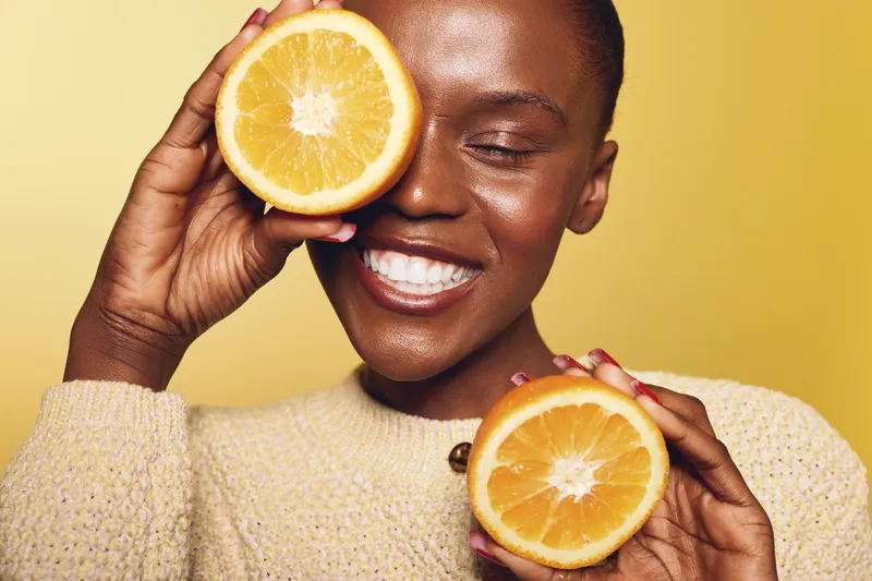 Smiling African woman with dark skin holds two halves of fresh orange near face on yellow background. Concept of healthy lifestyle, natural beauty, vitamin C, and happiness.