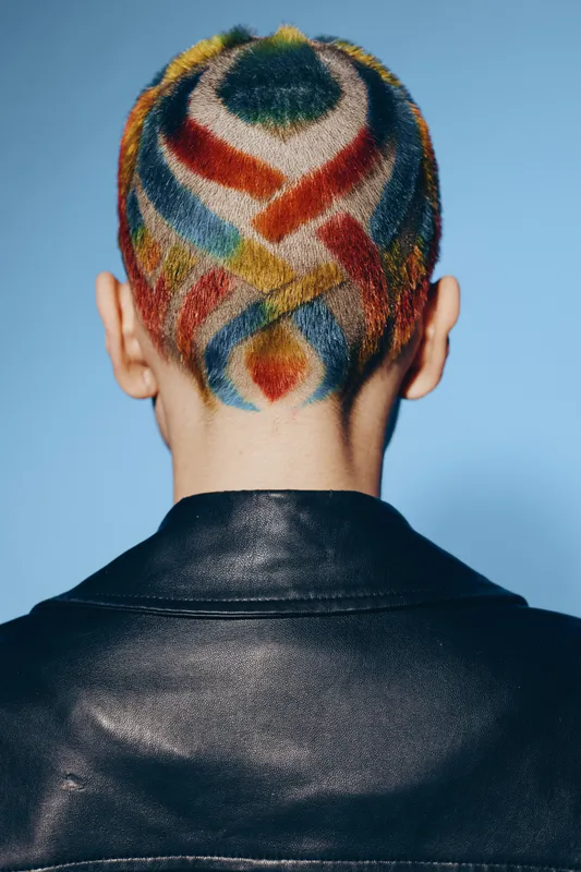 Creative woman with colorful shaved hair pattern and black leather jacket, viewed from behind against a plain blue background, showcasing modern edgy style.
