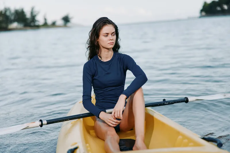 A solitary female rower sits in a small yellow boat on calm water, wearing a navy long sleeve shirt, poised with oars ready, focused gaze and calm demeanor.