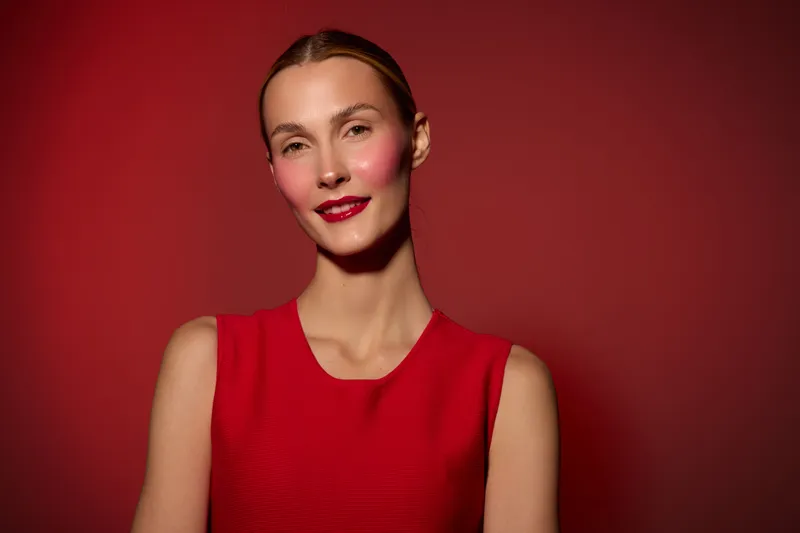 Portrait of a smiling woman with red lipstick and blush wearing a red sleeveless dress against a red background. Concept of beauty, fashion, and confident female model.