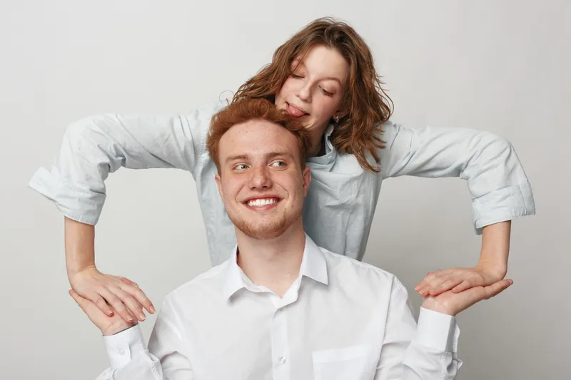 Happy young couple in white shirts smiling and posing together on grey background. Woman stands behind man holding his hands, showing affection and joy in studio.