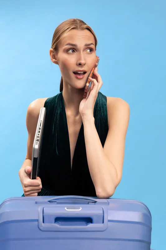 Young woman in black dress holds laptop and talks on cellphone with surprised expression. Blue suitcase in foreground, light blue background. Concept for travel, communication, and business trip.