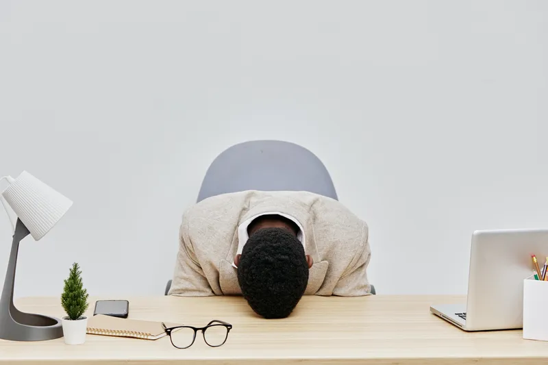 Professional young man with short curly hair wearing beige blazer leaning on desk head down in despair or stress at work in modern office environment. Concept: workplace stress or burnout.