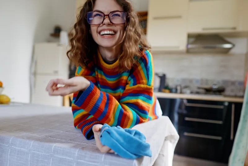 Cheerful woman in a colorful striped sweater cleans a table at home, smiling, showcasing a warm lifestyle moment in a cozy kitchen environment