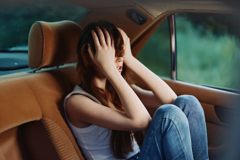A young woman experiencing emotional distress in a car, wearing casual attire, with a focus on her expressive facial features and subdued color palette.