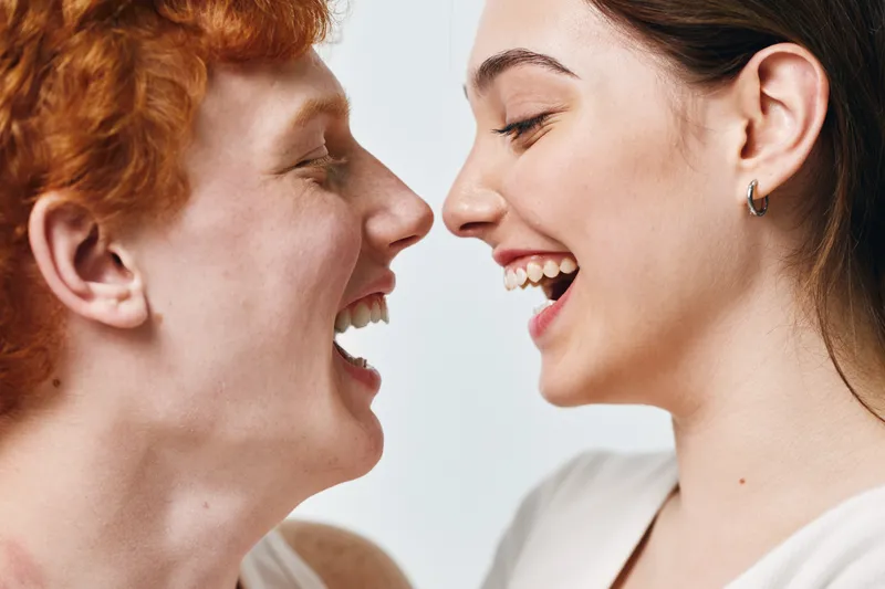 Two young smiling women facing each other with joyful expressions and open mouths, showing happiness and friendship on a light background in closeup portrait.