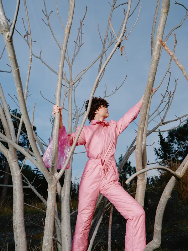 Outdoor climber in a pink jumpsuit navigates leafless trees under a clear sky, striking a dynamic pose that conveys movement, energy and playful strength in a natural setting