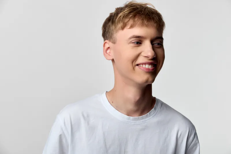 Smiling young man in a white t shirt, looking to the side with a friendly expression, casual lifestyle image highlighting positivity, approachable vibe, clean studio background, bright lighting