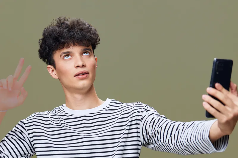 young man taking selfie wearing striped shirt making peace sign with hand against neutral background, casual style and relaxed mood