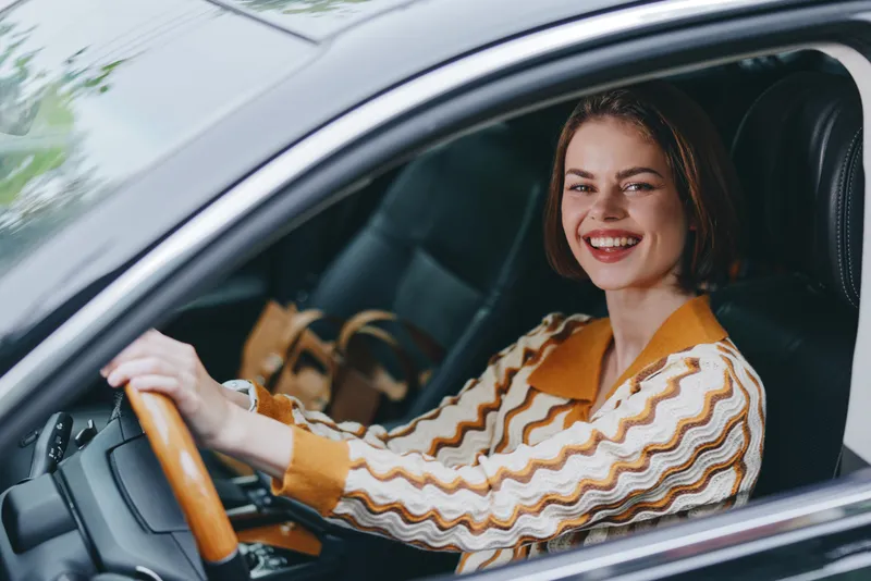 Smiling woman sits behind the wheel of a car, stylish striped jacket, warm lighting, and a confident, relaxed driver portrait inside the vehicle.