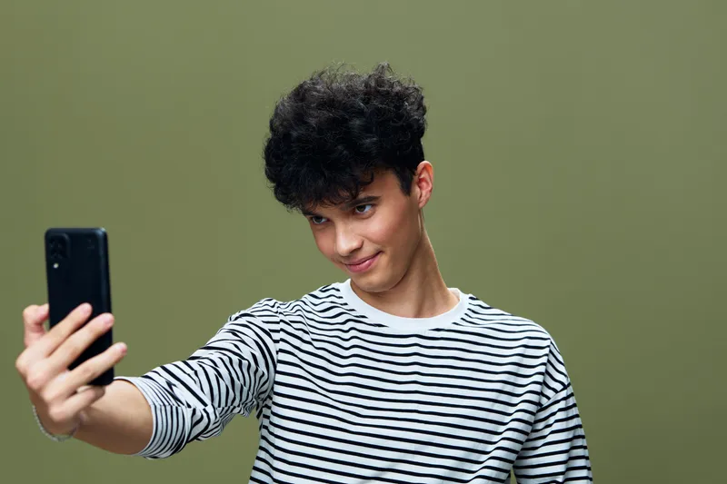Young man taking a selfie with a smartphone against a plain olive green background, wearing a striped shirt and smiling playfully at the camera.