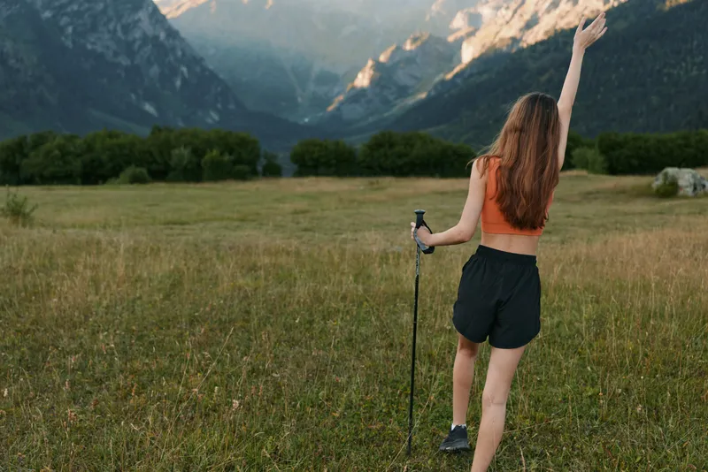 A lone hiker woman traverses a wide meadow framed by distant mountains. She wears a sleeveless top and shorts, raises one hand, and enjoys the fresh outdoor scenery.