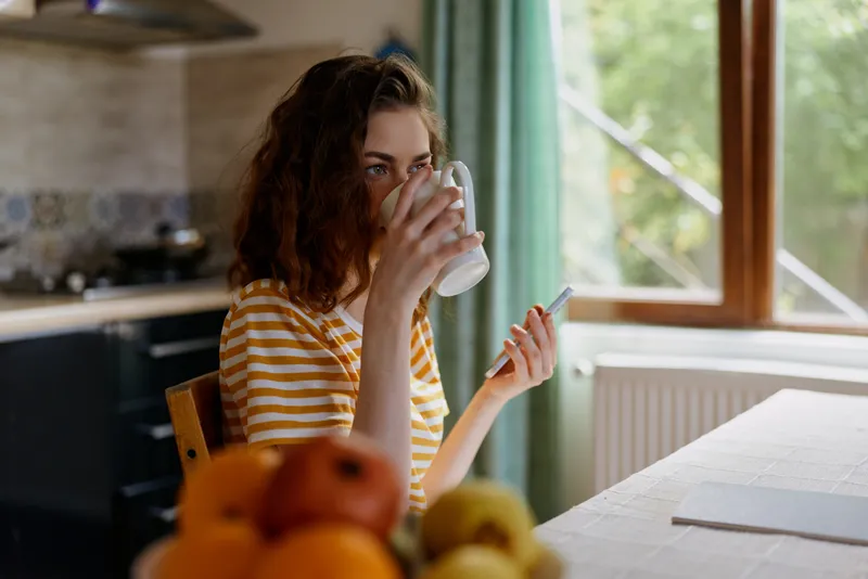 Woman enjoys a morning beverage at home in a bright kitchen, striped shirt, sipping from a mug, natural light, calm atmosphere, casual lifestyle scene, everyday cozy quiet