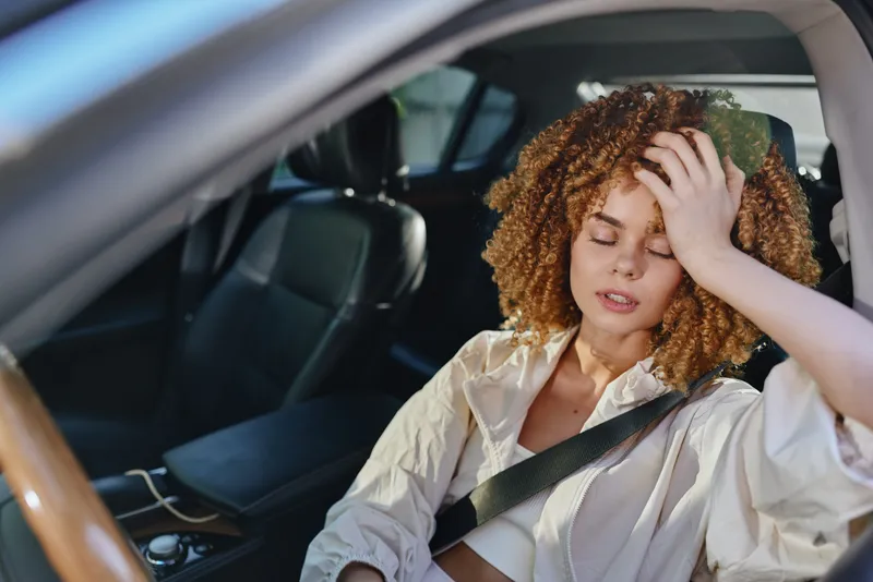 young woman feeling stressed sitting in car interior with leather seats, wearing casual light shirt and seatbelt, curly hair, daytime sunlight