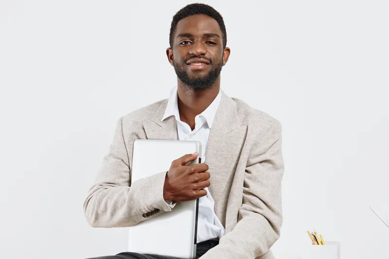 Confident smiling man holding closed laptop, dressed in beige blazer and white shirt, sitting against plain white background. Studio portrait of business professional, success, positivity, modern styl