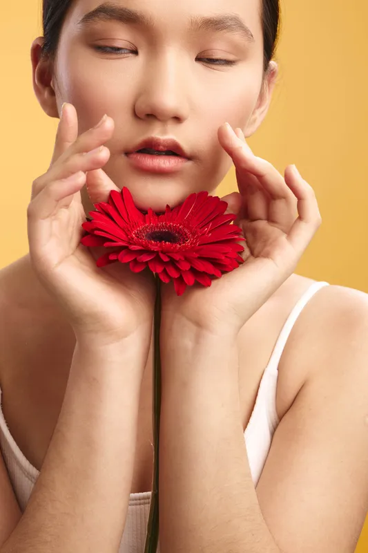Asian woman is captured in a soft closeup as she gently holds a vibrant red flower near her face. The serene expression and warm light create a graceful portrait.