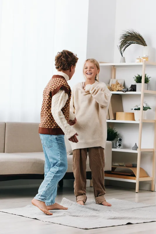 Two friends in cozy sweaters share a joyful moment indoors, dancing playfully in a bright living room with soft rug, natural light, and a calm home atmosphere.