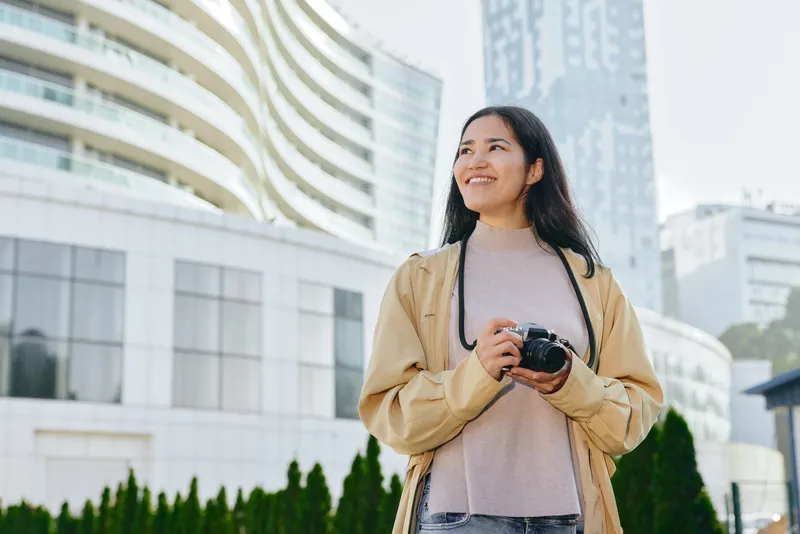 Camera in hand, woman stands outdoors in the city, capturing urban street scenes with glass towers, casual jacket, and a curious gaze toward the skyline.