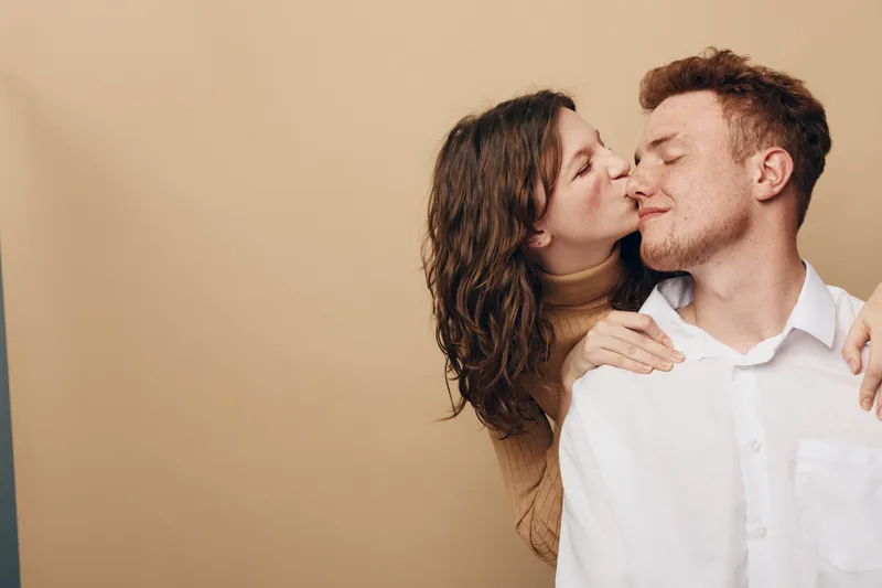 Young couple in casual clothes shares a tender moment, woman kissing man on cheek with closed eyes. Intimate love, affection, and romantic relationship concept on beige background.
