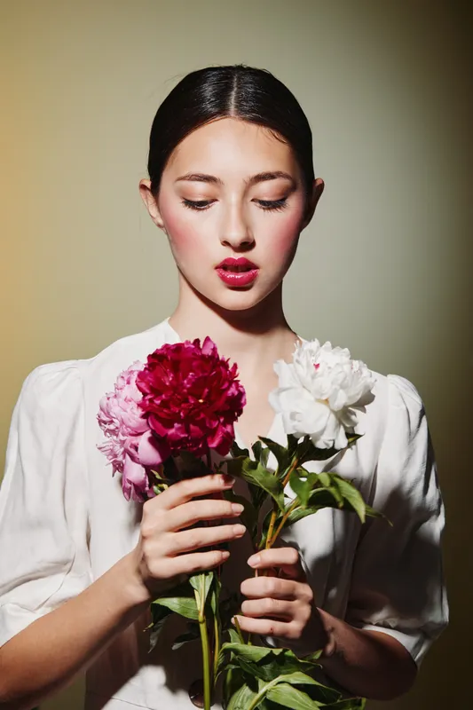 Young woman with slicked back hair holds bright pink and white flowers while wearing a white blouse. Clean portrait in soft light creates serene and stylish beauty concept.