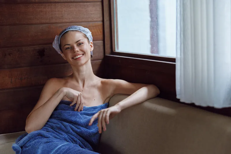 smiling woman wrapped in blue towel relaxing indoors near wooden window, natural light highlighting fresh complexion and calm mood after bath or spa session