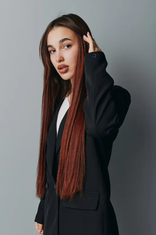 Young woman with long straight hair wearing black blazer posing against gray background. Portrait of confident female model with natural makeup and serious expression in studio light.