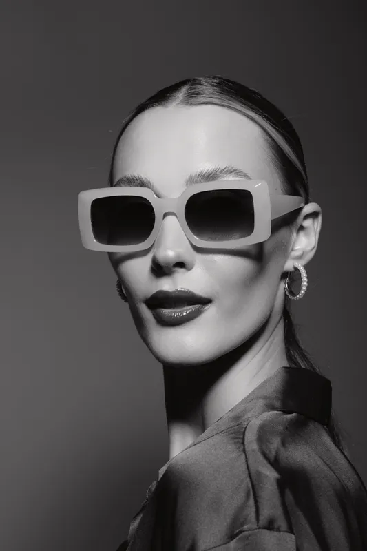 Elegant caucasian woman with pale skin wearing large sunglasses and hoop earrings poses confidently against dark background in black and white studio portrait.
