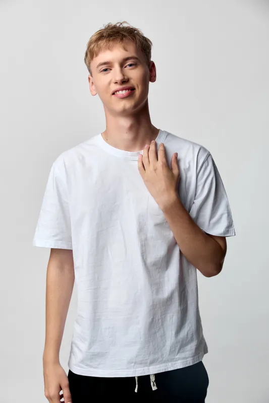Young man wearing a white t shirt in a casual pose, relaxed vibe, smiling softly in a clean studio setting, minimal wardrobe, everyday style, natural attitude, modern youthful look.