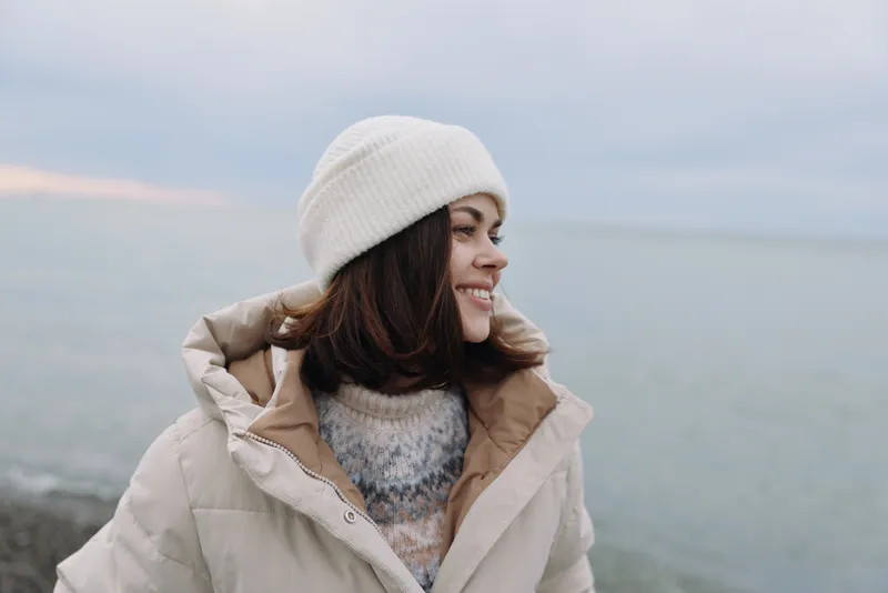 woman in a warm coat and beanie by the sea in winter, smiling softly as the coast breeze cools the air and calm waters reflect soft light on the horizon
