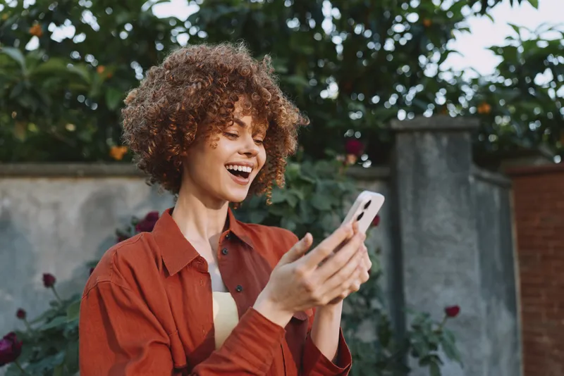 Smiling woman lifestyle outdoor portrait holding a phone, casual fashion, happiness, moment captured in a bright outdoor setting and relaxed ambiance