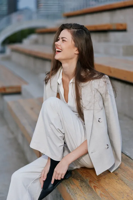 Woman laughing outdoors in stylish business outfit sitting on wooden steps with urban background, showing joyful emotion and casual confidence