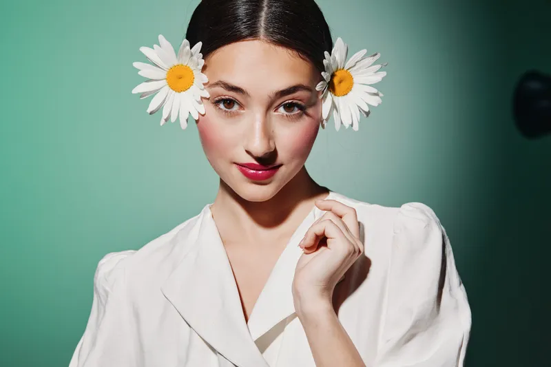 Young woman with fresh flowers in her hair poses confidently in a white blouse against a soft green background. Creative beauty and fashion concept with natural elements.