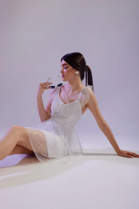 Elegant woman in a white lace dress sits on the floor holding sunglasses. Soft lighting highlights her beauty and fashion style in a minimalistic studio setting. Concept for elegance.