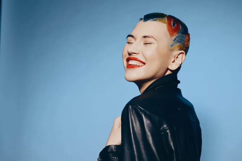smiling woman with colorful short hair wearing black leather jacket, expressing happiness against a plain blue background, joyful urban portrait style.