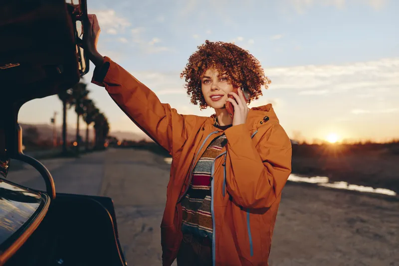 Woman lifestyle portrait by car at sunset, curly hair, wearing orange jacket, smiling while speaking on a smartphone, one hand to hair, urban road and evening sky, relaxed mood