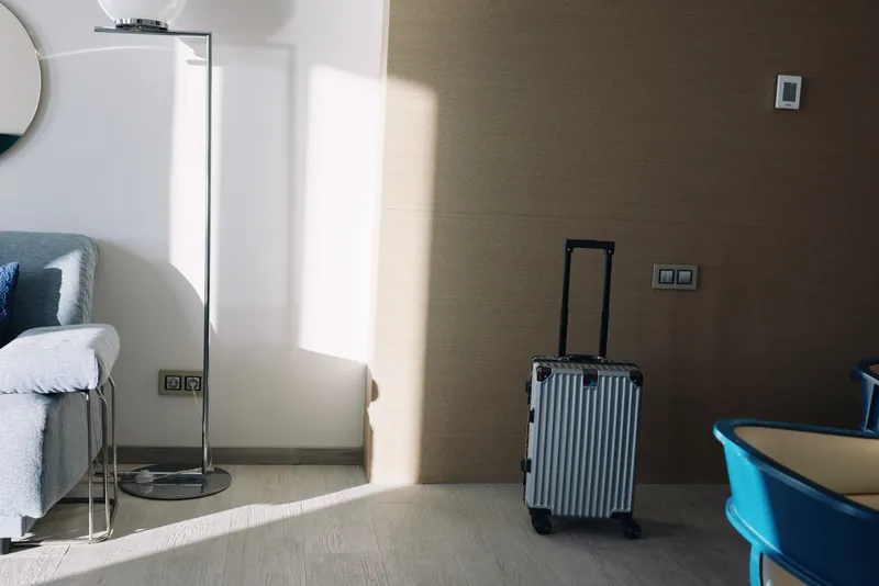 Luggage and suitcase by a sunlit hotel room, showcasing travel vibe, modern interior, bed and lamp with a blue chair in calm morning light and soft shadows.