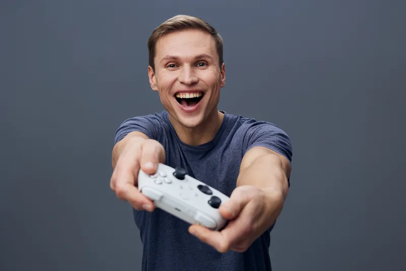 Enthusiastic young man holding game controller, smiling broadly, celebrating victory, casual t-shirt, short brown hair, isolated on plain gray background, studio portrait. People lifestyle concept