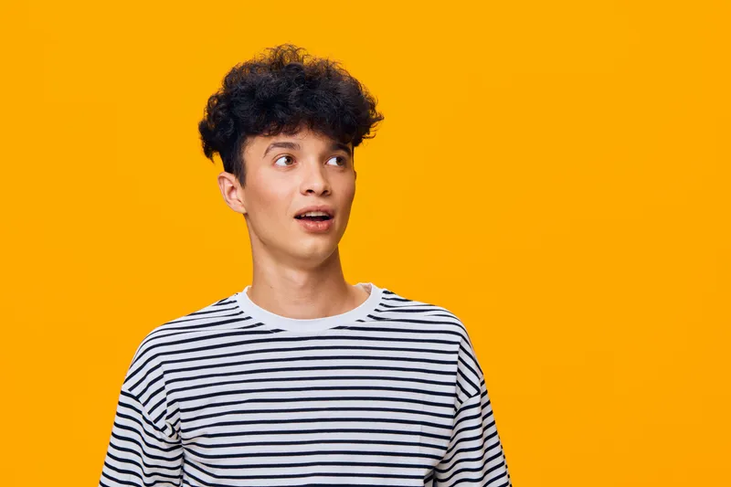 young man with curly hair wearing striped shirt looking surprised against bright orange background, casual style, expressive emotion, studio portrait