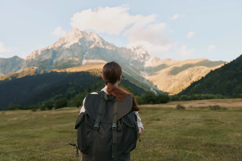 Woman traveler with backpack stands in grassy field looking at mountain landscape under blue sky with clouds, enjoying outdoor adventure and nature exploration.