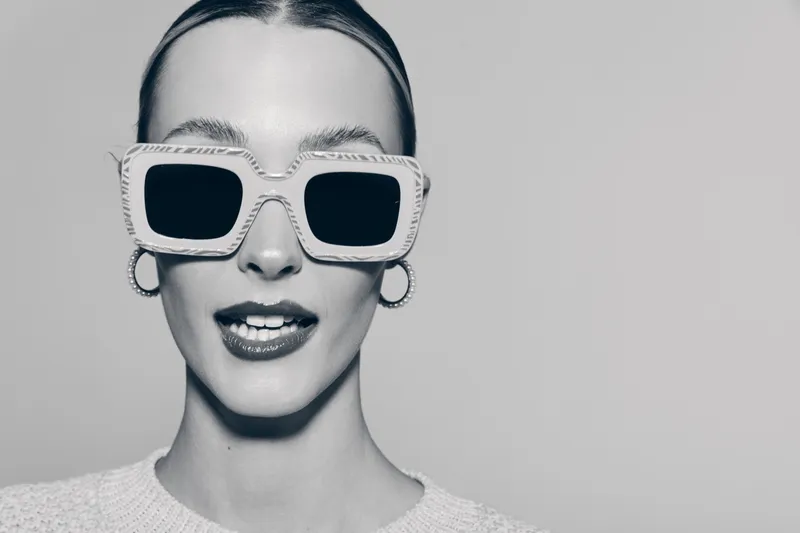 Young white woman with pale skin wearing large sunglasses and hoop earrings sticks out tongue, studio portrait in black and white.