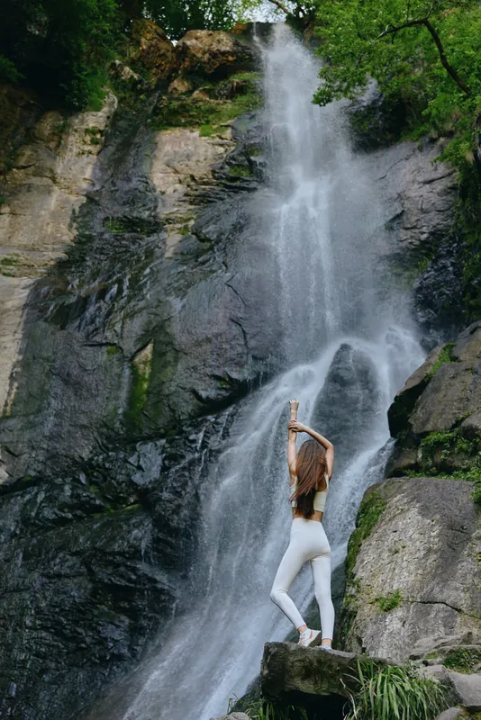 woman standing near waterfall, natural rocks, flowing water, green trees, outdoor adventure, white outfit, peaceful nature scene