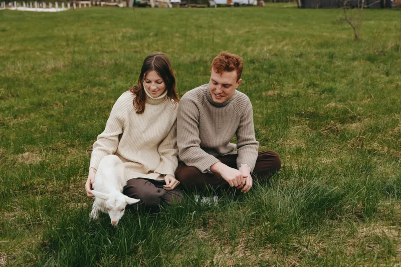 Couple sits on green grass outdoors as a playful goat kid approaches, capturing a warm, relaxed lifestyle moment in nature and gentle companionship